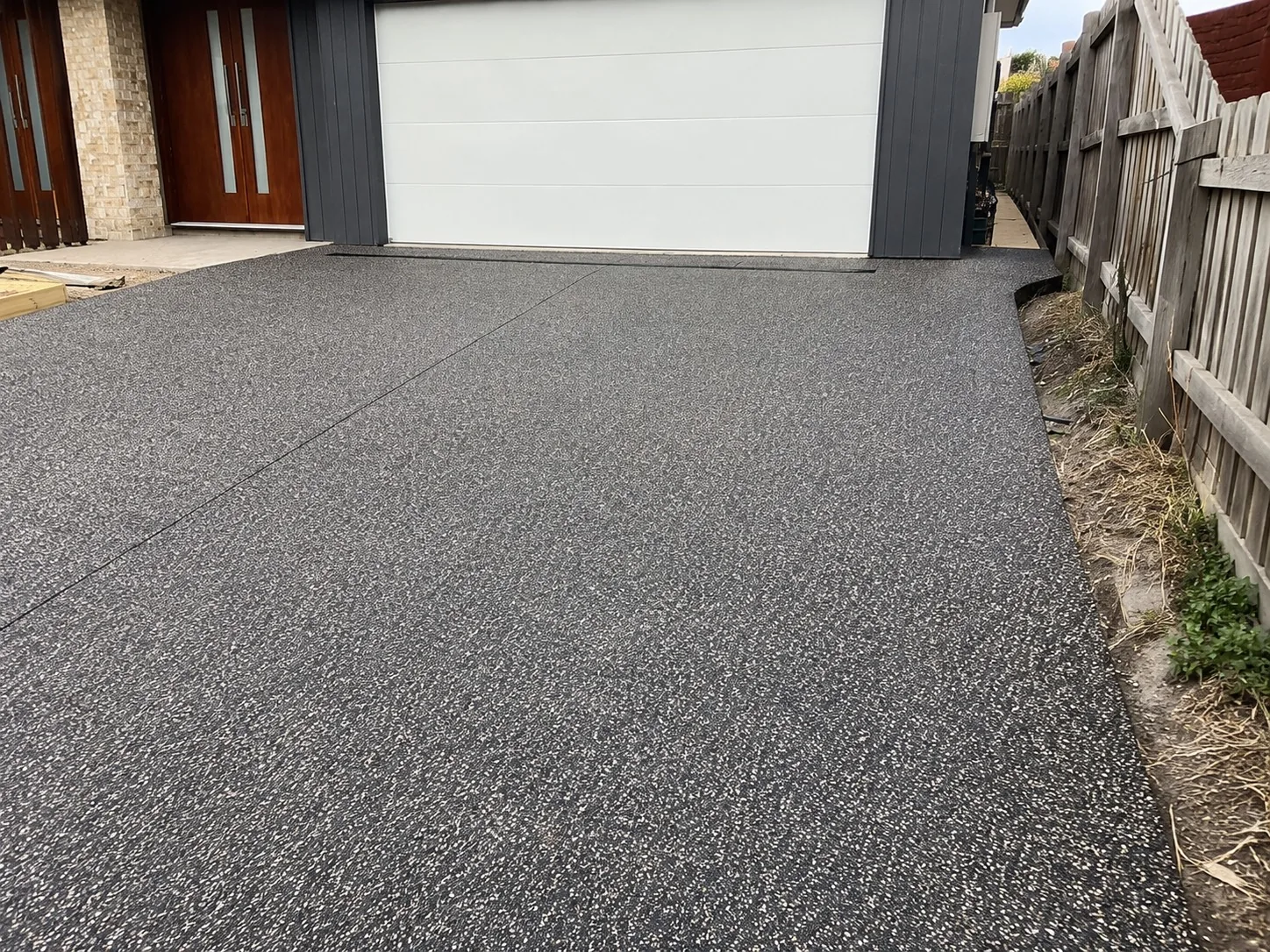 Charcoal exposed aggregate driveway running between a timber fence and a white garage door