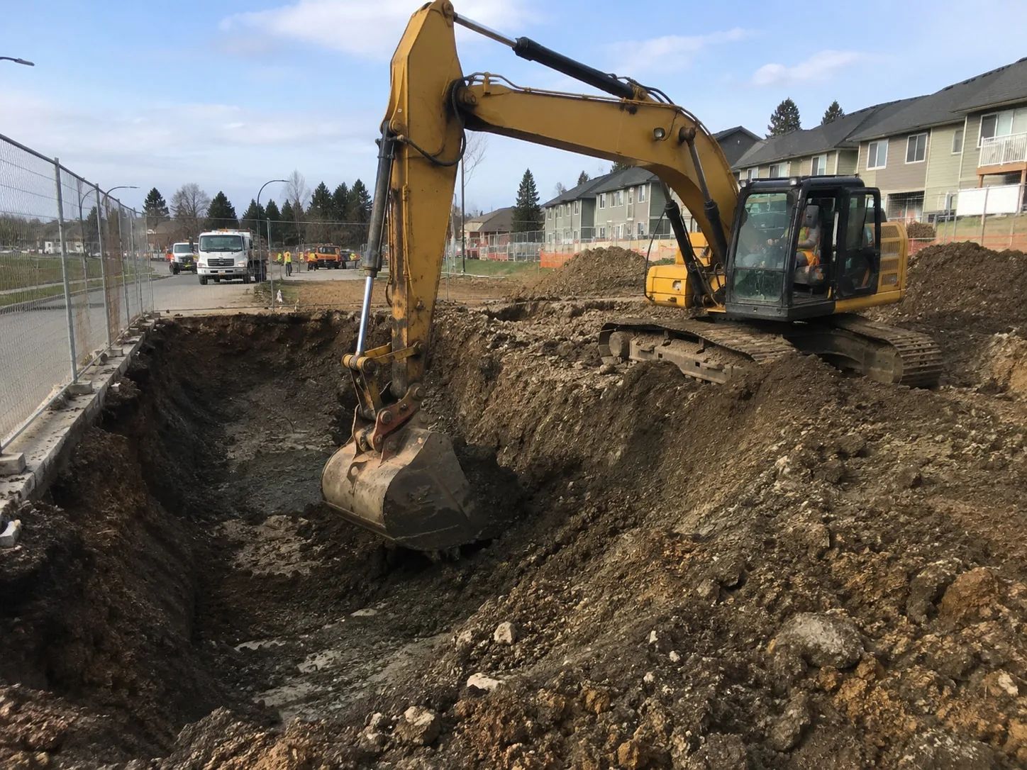 Excavator digging a deep excavation beside a residential street with spoil stockpiled alongside