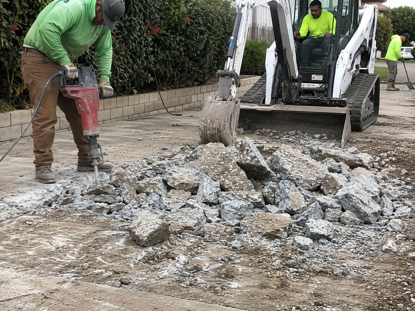 Worker breaking up a concrete slab with a jackhammer beside broken concrete and a mini excavator