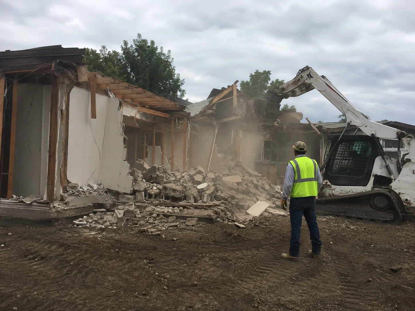 Machine demolishing a small building while a worker in hi-vis clears rubble on site